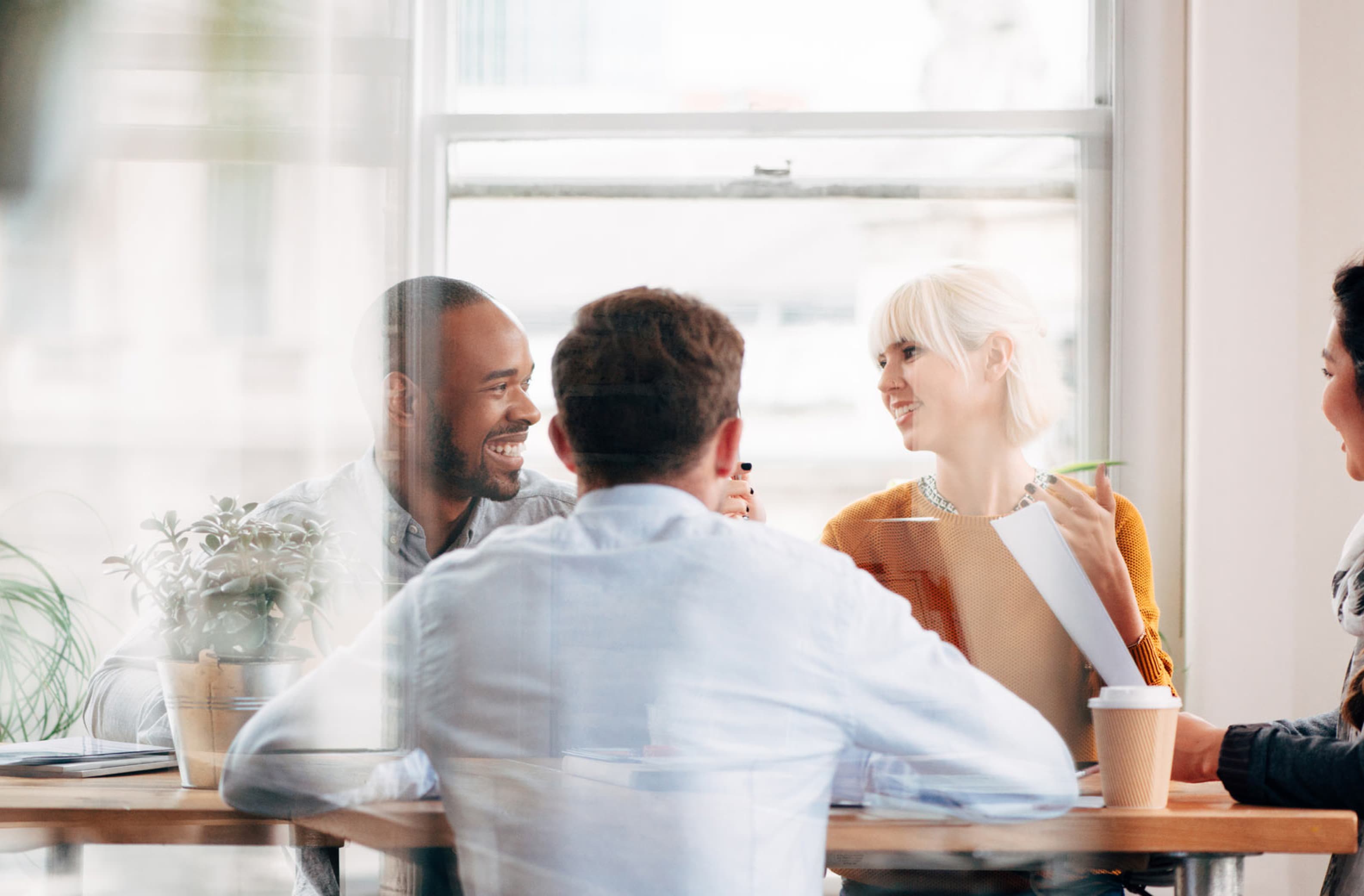 Vier Personen im Gespräch am Tisch, lockere Atmosphäre mit Pflanzen und Kaffeebecher im Büro.