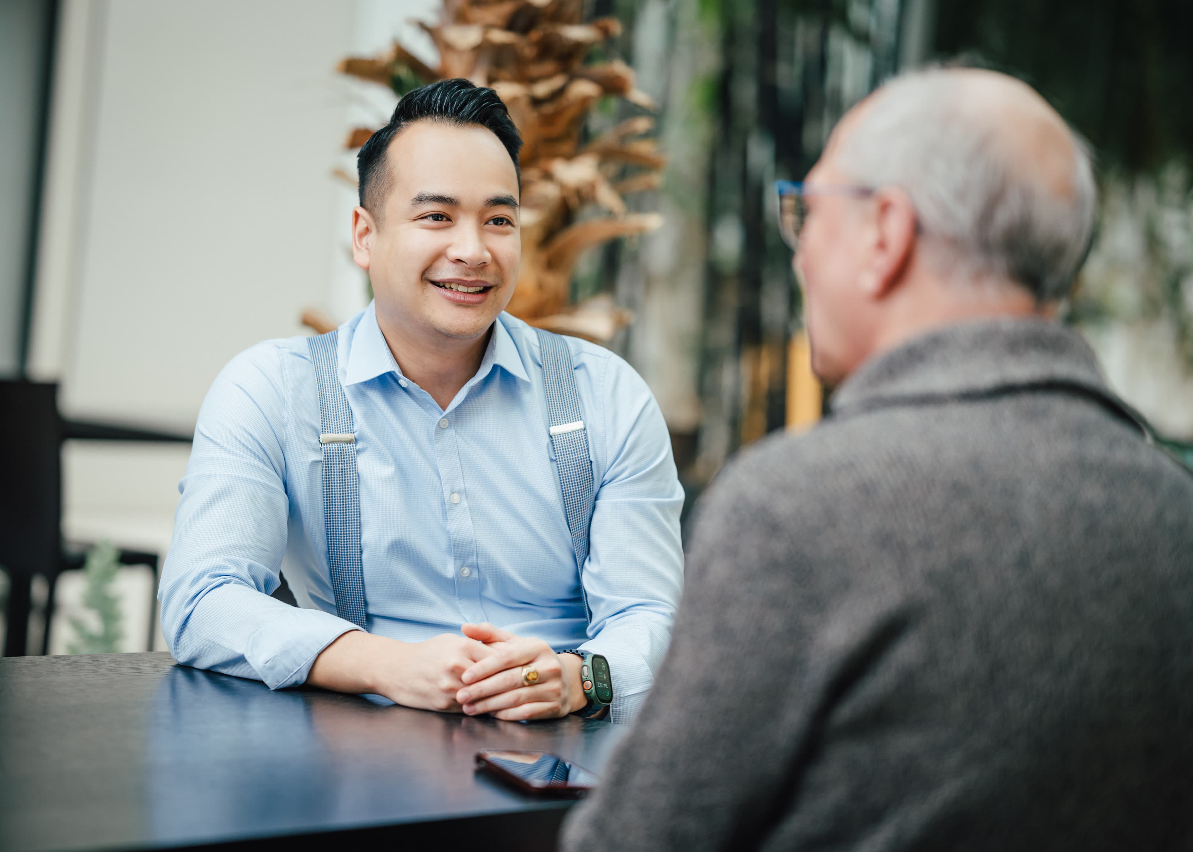 Zwei Männer sitzen an einem Tisch und sprechen miteinander, einer trägt Hemd mit Hosenträgern, der andere grauen Pullover.