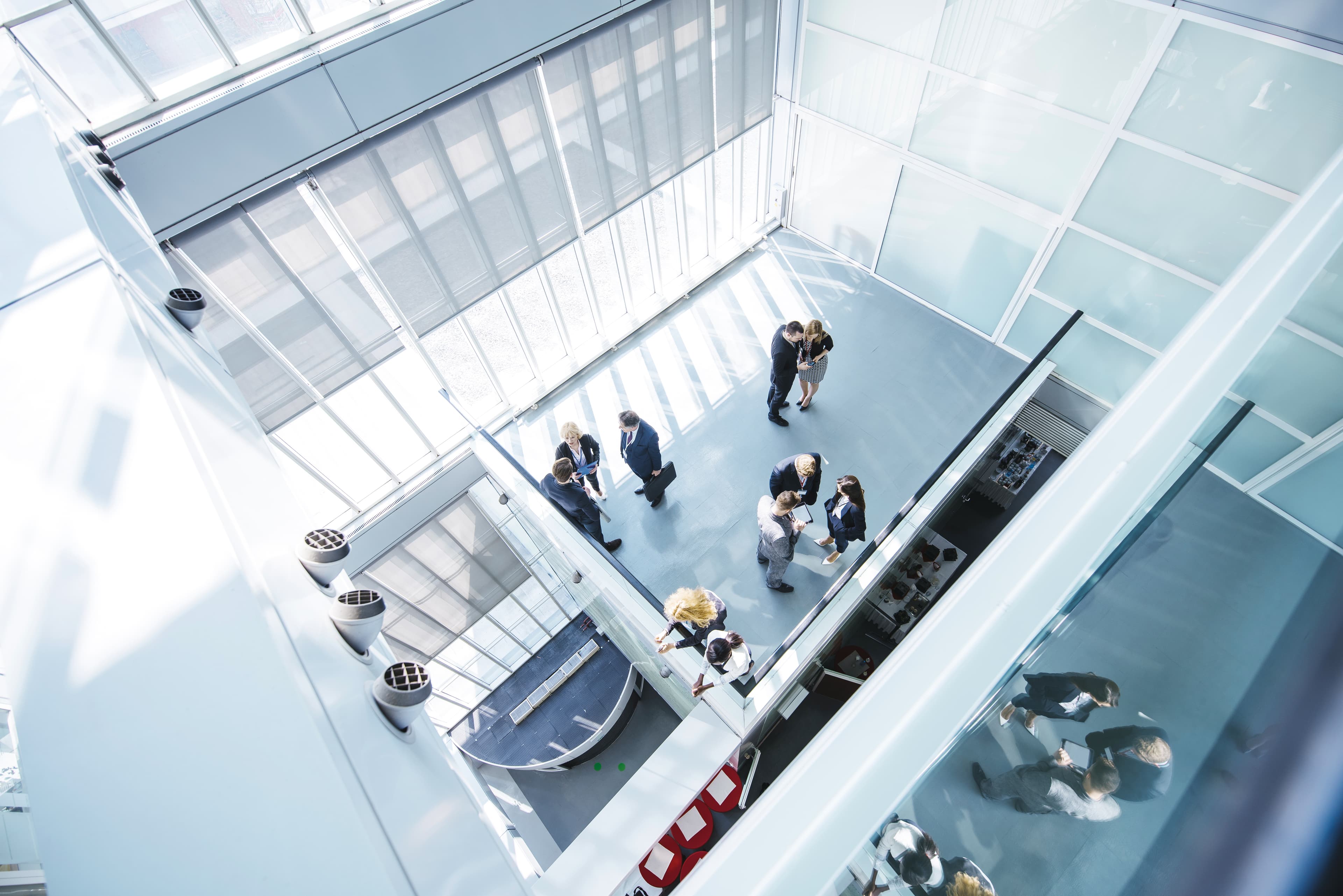 Blick von oben auf eine Gruppe von Menschen in Businesskleidung, die sich in einem modernen, lichtdurchfluteten Atrium unterhalten. Das Glas der Architektur spiegelt einige der Personen.