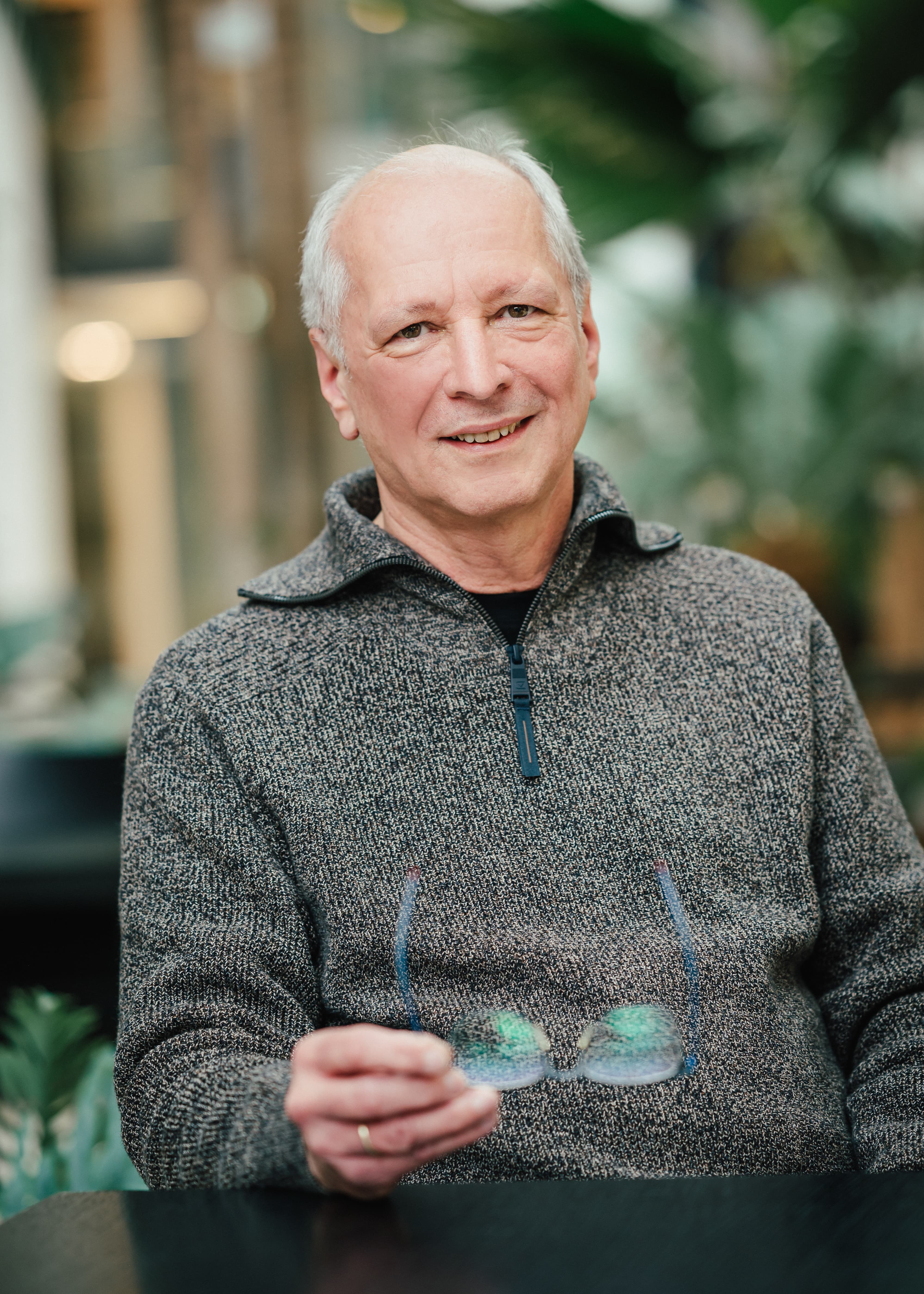 Ein älterer Mann im grauen Pullover sitzt lächelnd am Tisch und hält seine Brille in der Hand.