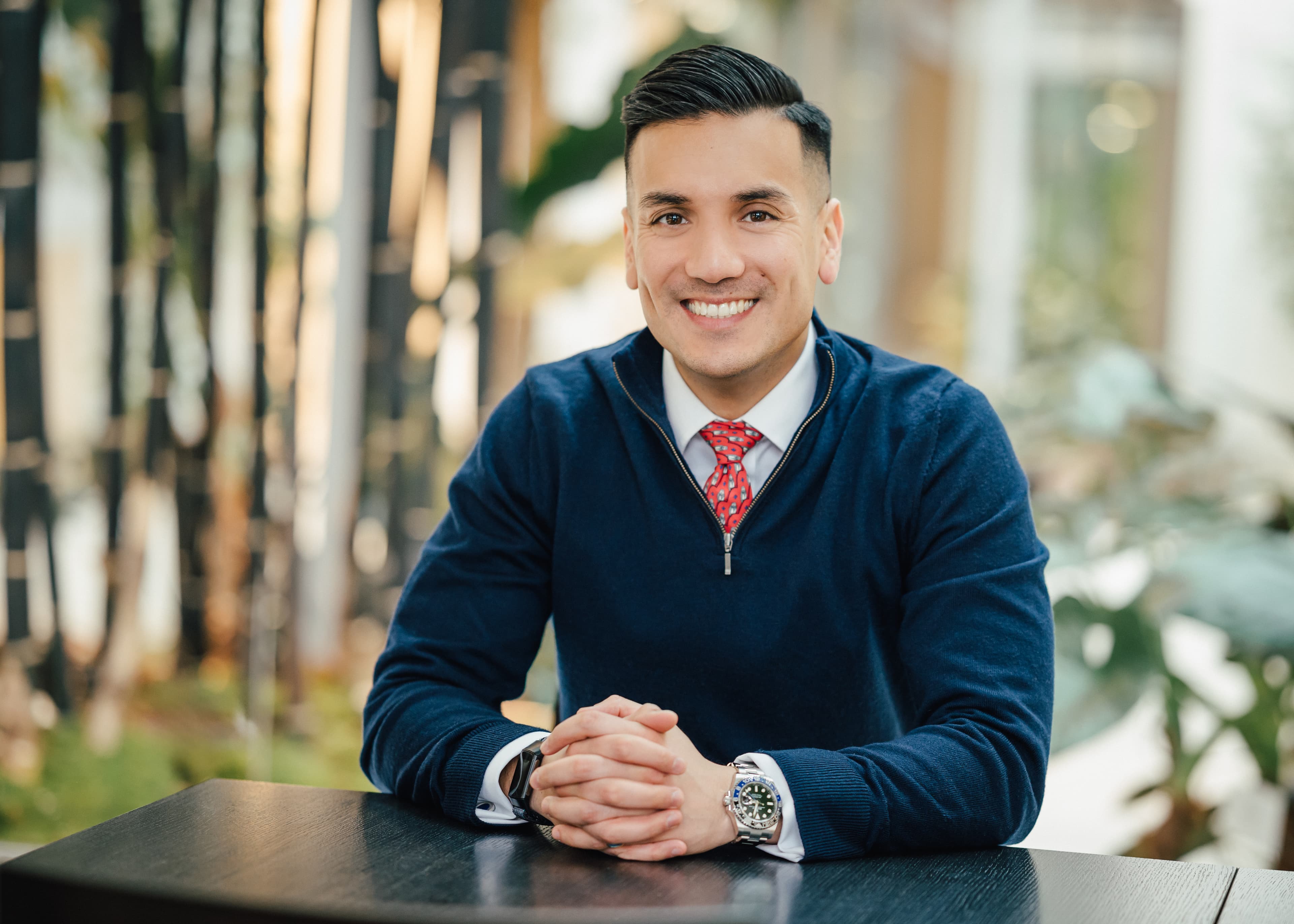 Porträt eines Mannes mit dunklen Haaren, verschränkten Händen und freundlichem Lächeln.