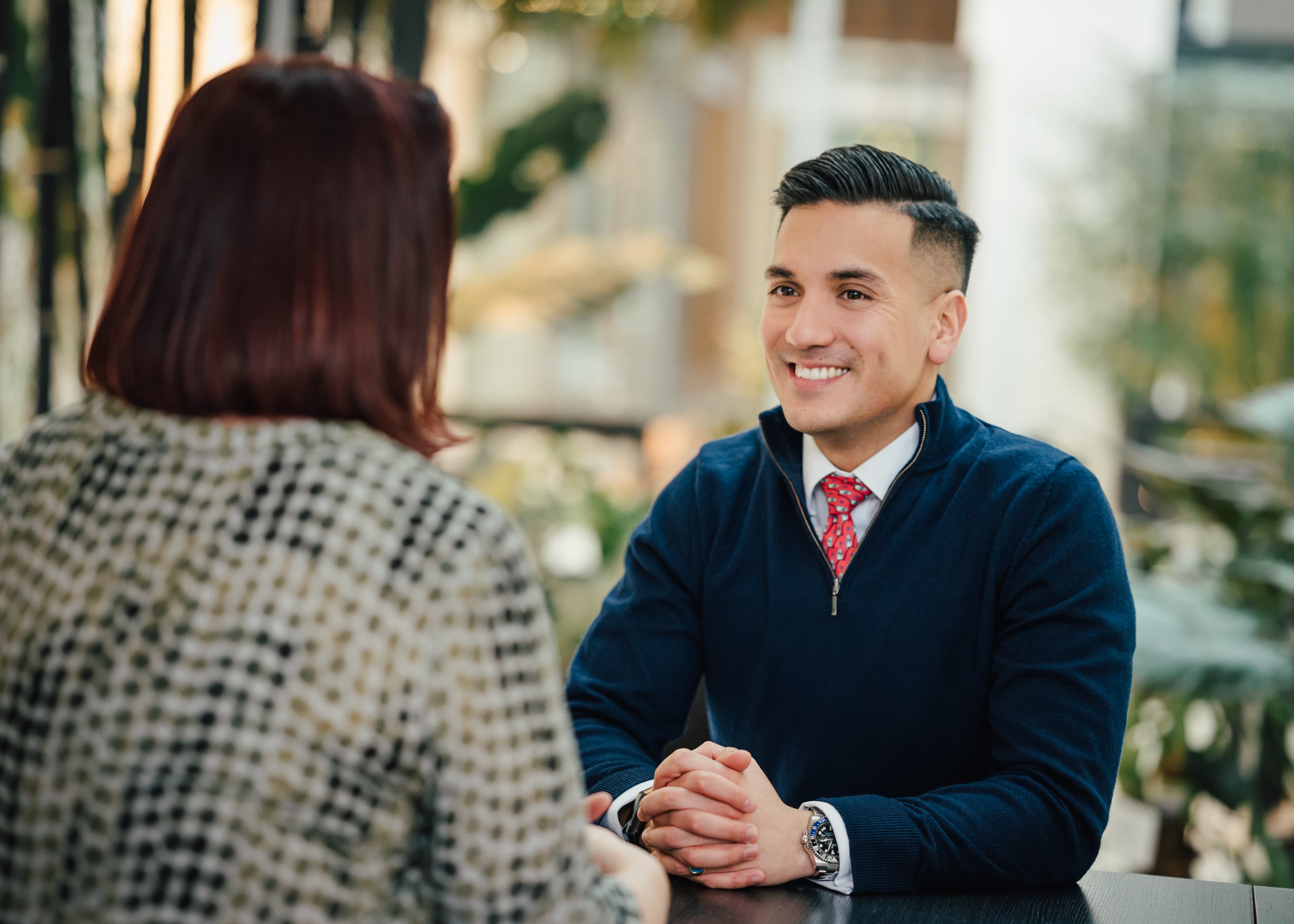 Ein Mann im blauen Pullover sitzt lächelnd einer Person gegenüber, beide im Gespräch bei der Baader Bank.