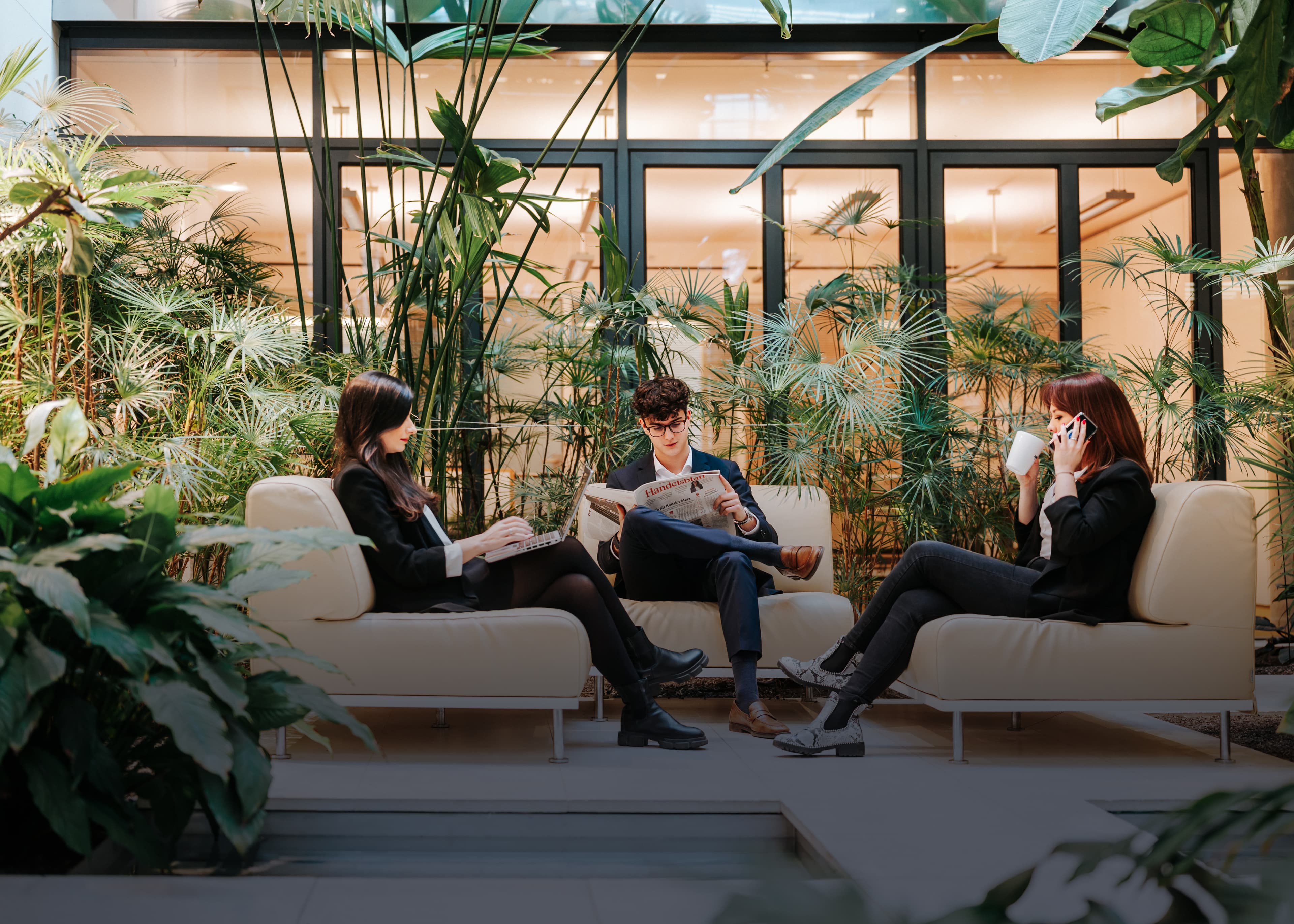 Drei Mitarbeitende der Baaderbank sitzen in einer begrünten Lounge mit Zeitung, Laptop und Kaffeetasse.
