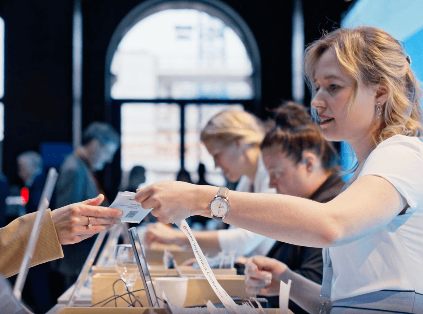 Mitarbeiterin der Baaderbank übergibt ein Ticket an eine Person am Empfangstisch.