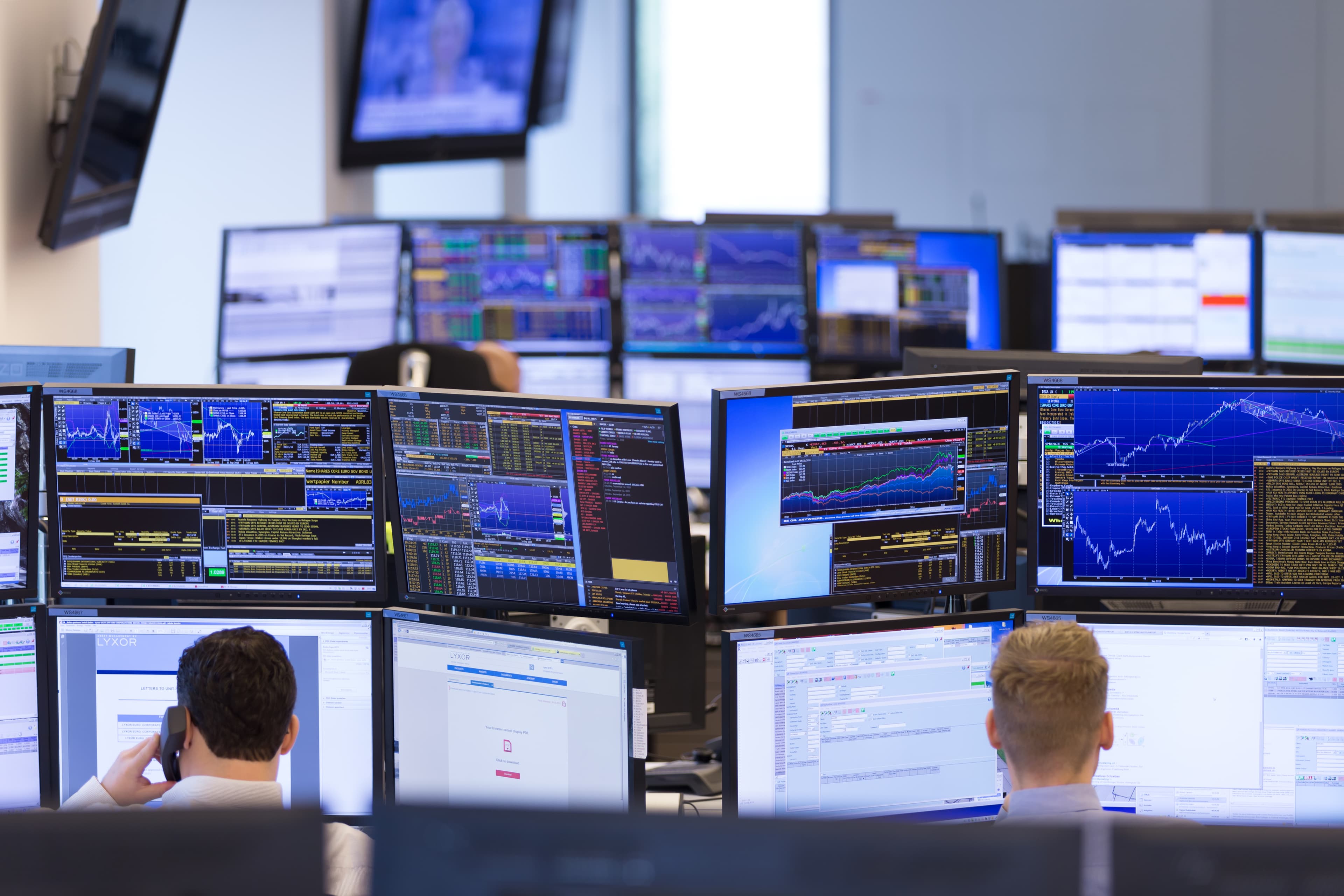 Arbeitsplätze mit vielen Monitoren, auf den Bildschirmen sind Finanzdaten und Kursgrafiken zu sehen.