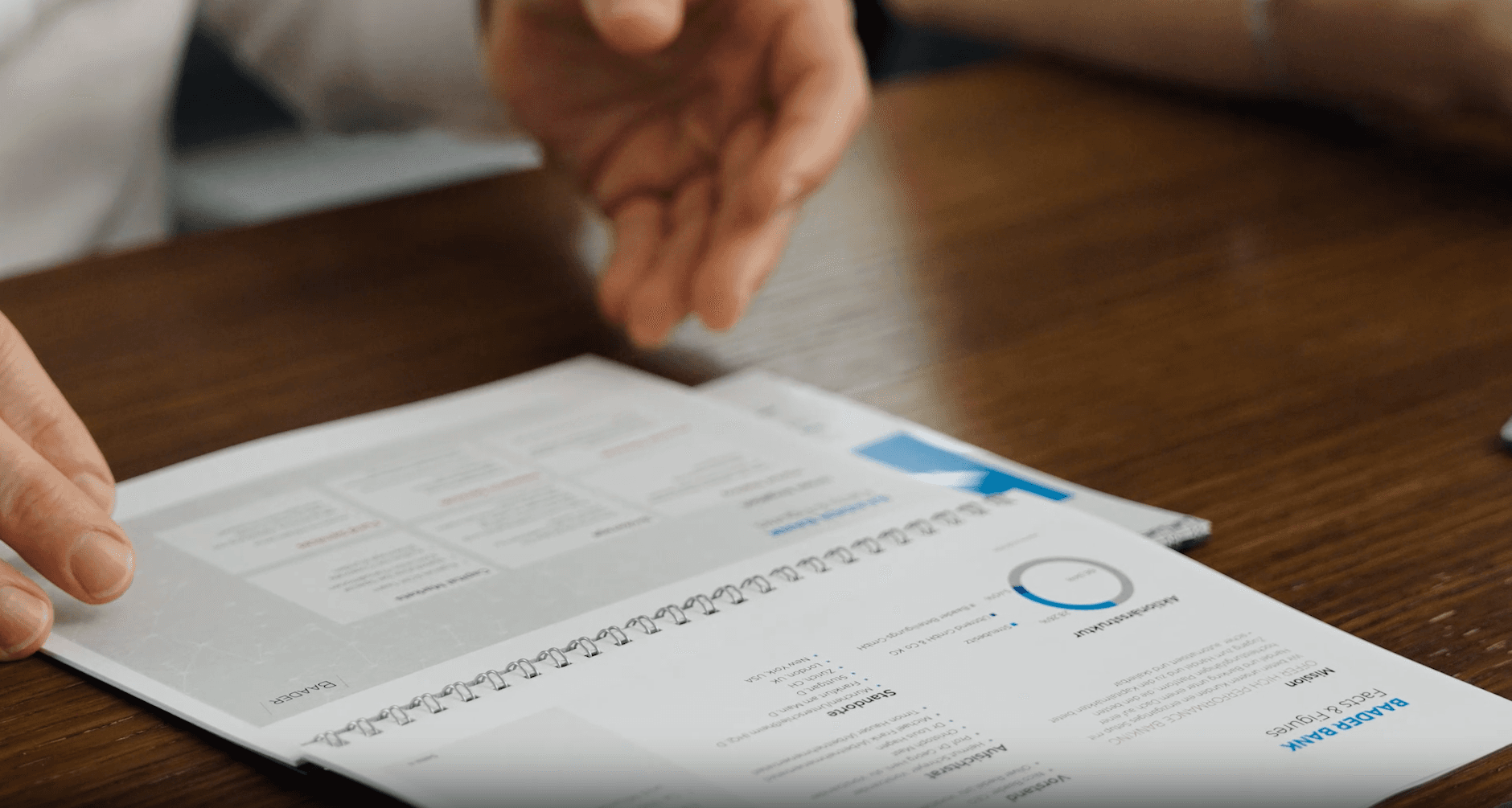 uf einem Holztisch liegt ein aufgeschlagenes Ringbuch mit dem Titel „Baader Bank Facts & Figures“, daneben eine ausgestreckte Hand.