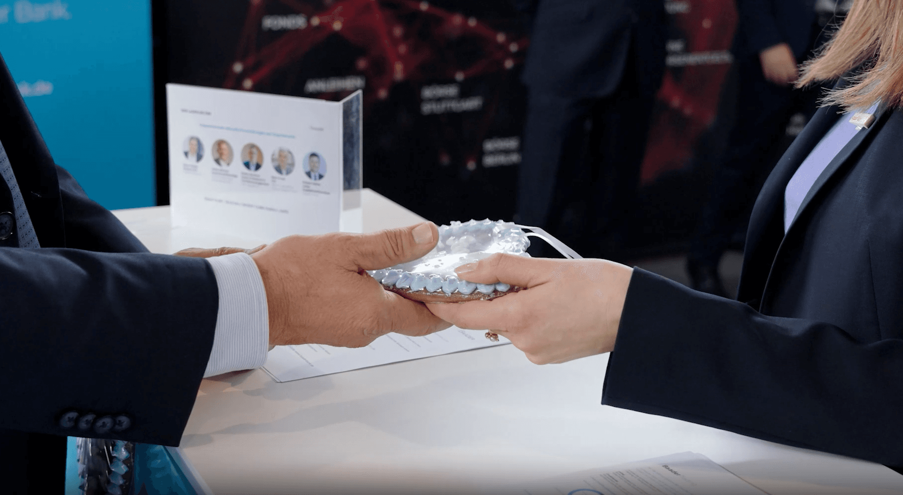 Zwei Personen im Anzug reichen sich ein Lebkuchenherz an einem Messestand.