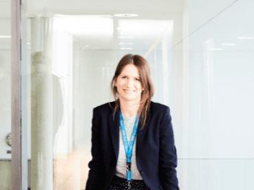 Frau mit dunkler Jacke, weißem Oberteil und blauem Namensschildband steht in einem lichtdurchfluteten Büroflur mit Glaswänden, lächelnd in die Kamera blickend.